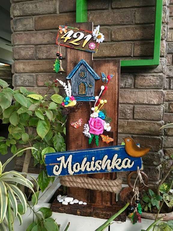 Mountain Hut Wooden Name plate
 As the name suggests, The design is inspired from huts made on high mountains, where nature is around and Birds make their nests in the mountains. We have used natural wood for the base and decorated it with artificial flowers, MDF cut bird and hut, wooden beads and hangings. The name is specially designed with small beads, please zoom and see the beautiful bead work on the name. The Jute string is beautifully places under name plaque. We have given space for address on the top. 